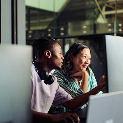Two colleagues working together at a computer in an office with large windows, engaged in discussion.