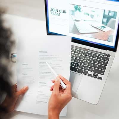 Person reviewing CV with pen while viewing job recruitment website displaying 'Join Our Team' on laptop screen