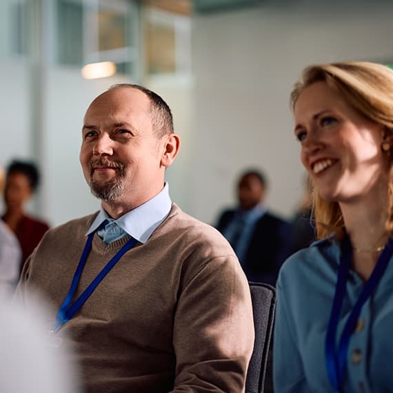 Two professionals with blue lanyards smiling during a presentation, man in beige sweater and woman in teal jacket.