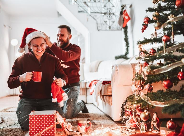 Smiling Gay Couple Opening Christmas Presents With Santa Hats And Drinking Tea Together