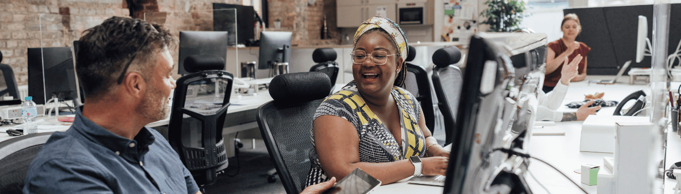 Smiling woman in colorful attire sitting at desk with coworker in modern office with computers.