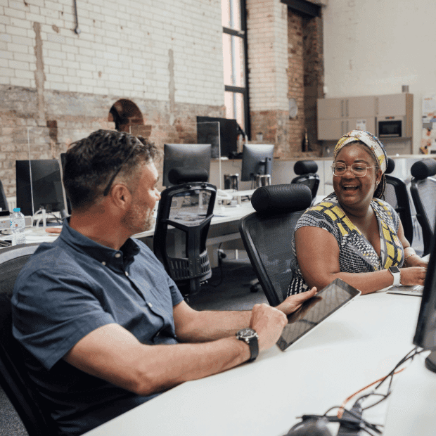 Two coworkers smiling and conversing at desks in a modern office with computer monitors.